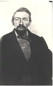 Portrait of Ash Upson, seated wearing a dark suit, sweater and bow tie