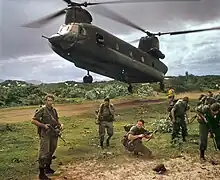 Coloour photo of a group of men wearing military uniform and carrying guns standing in front of a twin-rotor helicopter which is flying just above the ground