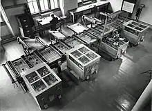 Looking down from above at a room at an intricate mechanical device which fills the room. In the background, a man sits at a desk next to a filing cabinet.Owens 1991, p.&nbsp;15.
