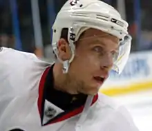 A head shot of a Caucasian hockey player wearing a white helmet with a visor and a white jersey