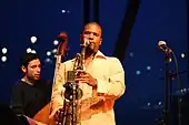 A black man in the front wearing a light orange shirt and playing the saxophone with his eyes closed and in the back a white man with his eyes closed playing the cello.