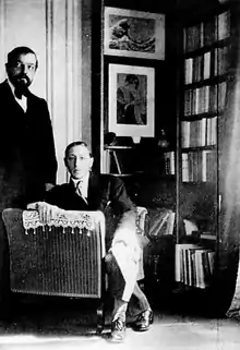 two white men, one bearded, middle-aged, standing, one younger, seated, in a book-lined room