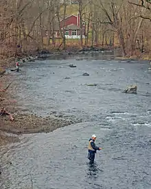 A wide muddy stream with some rapids surrounded by woods with bare trees and a red structure visible through them behind a bend at the rear. At the left are two men knee-deep in the water, holding fishing rods with a colored line attached