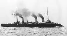 A large warship with two large guns steams at high speed, with thick black smoke pouring from its three funnels.