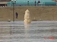 Water from a river completely surrounds a stone obelisk. In the background are several people with news cameras. A building with a green roof is seen behind them. At the bottom right of the image is the camera's time: "6 7:59 am".