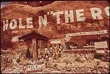 very large painted sign on the side of a sandstone cliff reading Hole N" The Rock with arrows pointing to the entrance. A handful of visitors are visible walking towards the door. Multiple signs are visible as well as a small rock garden.