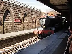 A railway vehicle disguised as a red-colored steam locomotive pulling into an old-fashioned railroad station building.
