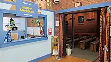 On the left, a sign reads, “Welcome to Puppet Showplace BOX OFFICE.” Below is a light blue wall with an opening and counter. Puppets are visible on the counter, through the opening, and near the sign. On the right, an opening in a brick wall leads to the theater. Colorful curtains frame the opening. In the theater, there are benches with light blue cushions.