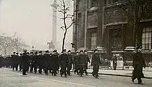 Black-and-white photo of a large group of men wearing military uniforms marching in formation along a city street