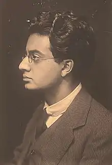 A man in his mid-20s. He is wearing a suit and a tie and is looking to his right