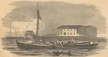 a black and white image of a wooden steamboat lying low in the water near a shore with buildings. The smokestack of the steam engine lies flat in a mass of broken planks in the center of the boat while men clamber about the wreckage. Three men in a rowboat observe the scene.