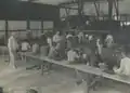 Europeans (standing in white suits) speaking to plantation workers (primarily sitting), c. 1905